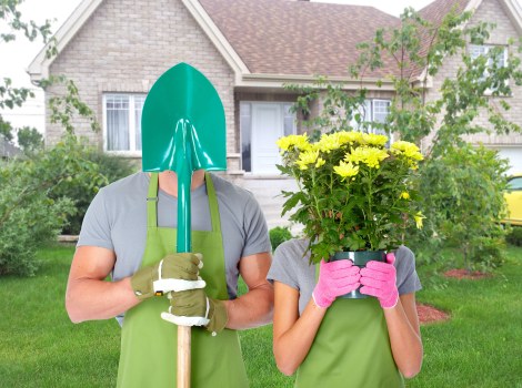 A man and woman in outdoor garden attire, both wearing green gardening aprons, stand on a well-maintained front lawn of a residential property in Highgate. The man, wearing a grey shirt, holds a teal, plastic garden shovel vertically in front of him, partially hiding his face with the shovel's handle. The woman, dressed in a grey t-shirt, is holding a potted plant with vibrant yellow flowers close to her face, obscuring her features. Behind them, there is a neatly landscaped garden with a lush green lawn, a variety of shrubs, and a young tree with light green foliage. The background features a light-colored brick house with white-framed windows and a pitched roof, set in a suburban neighbourhood typical of Highgate. The scene appears to be on a bright, slightly overcast day, with soft natural light illuminating the outdoor space, clearly showing the garden's elements and the gardening activity as part of professional outdoor maintenance or planting services by Gardening Services Highgate.
